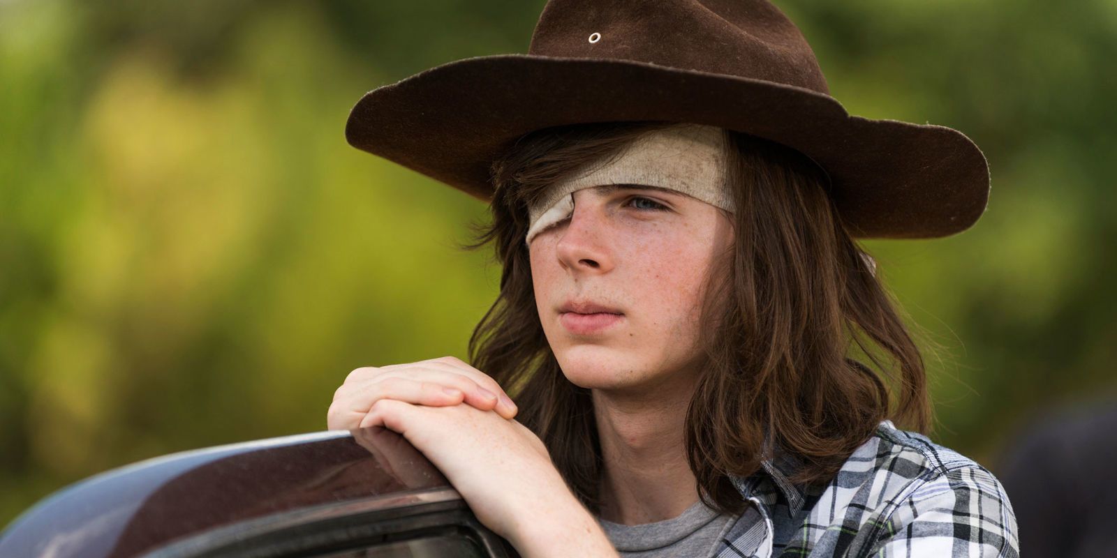 Carl Grimes leans on the door of a car while wearing an eyepatch and hat