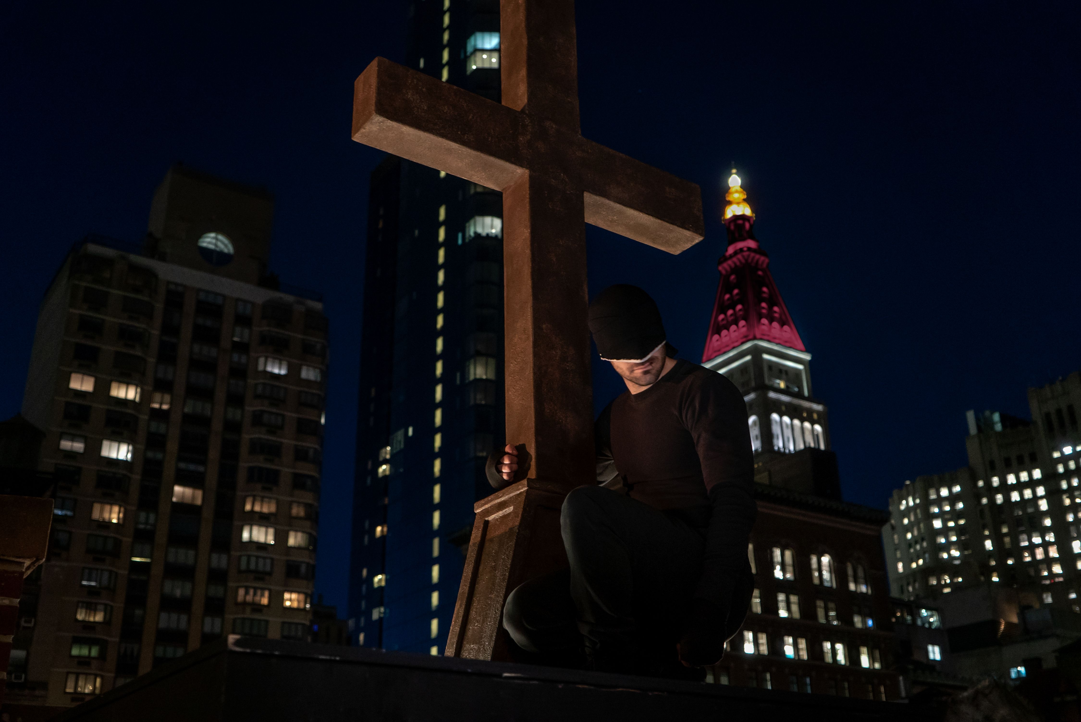 Daredevil drapes his arm around a cross in Season 3 photo.