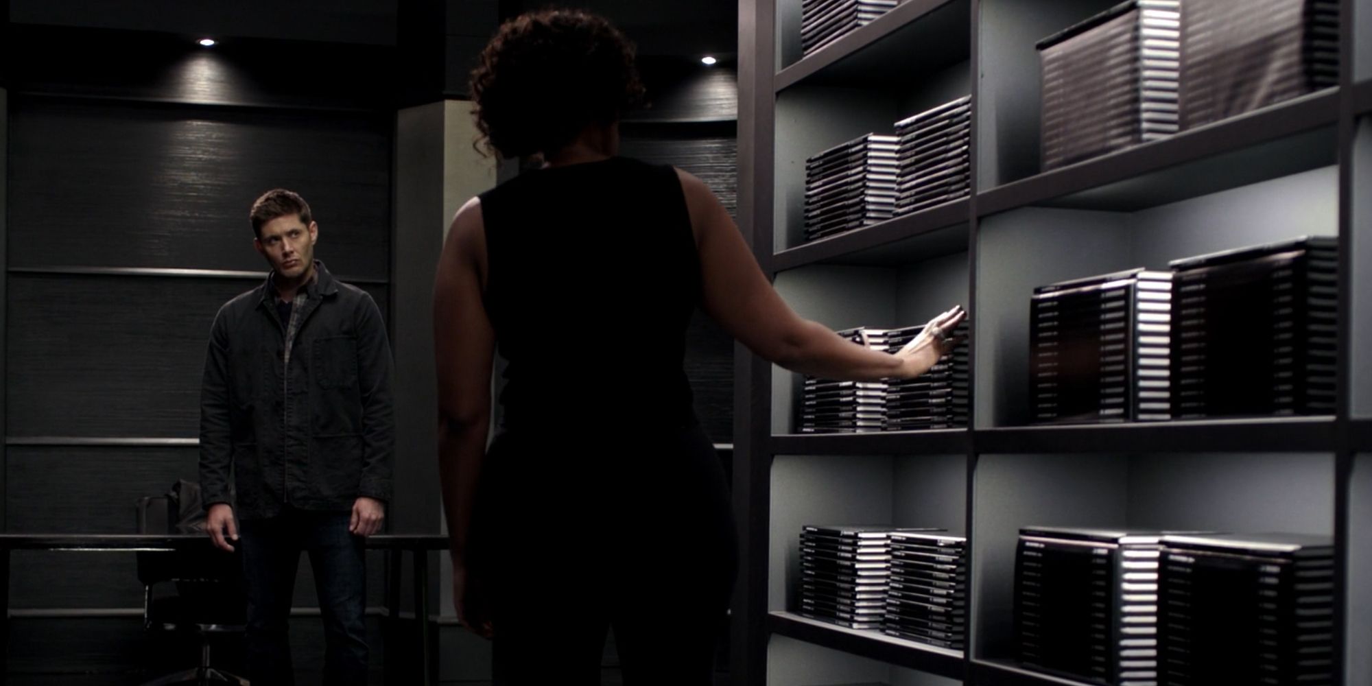 Dean standing in front of Billie (Death) and a shelf full of books.