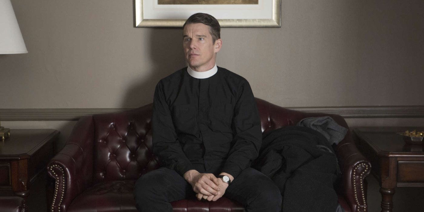 A young priest sits on a couch, hands clasped in his lap.