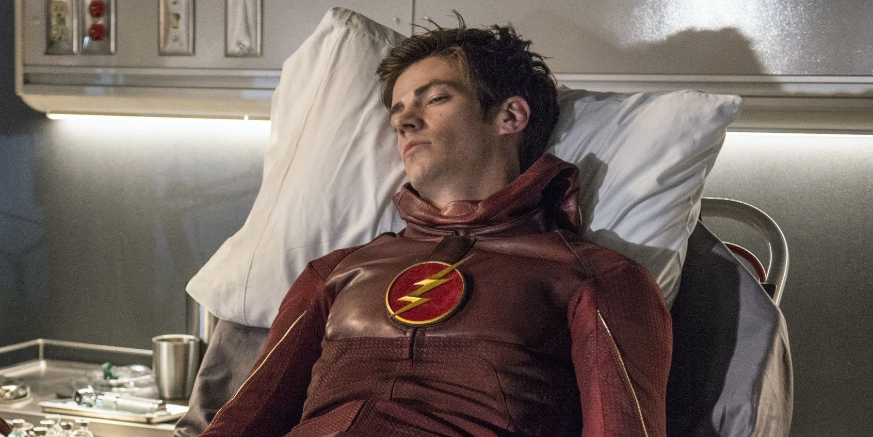 Barry Allen lying in a hospital bed, looking beat up and with his suit unzipped from the neck up to reveal his face