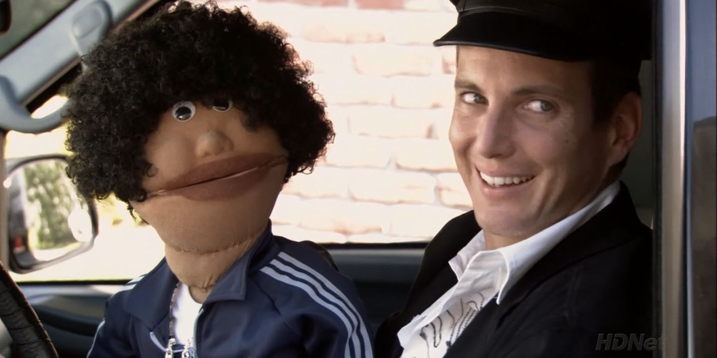 Gob from Arrested Development sitting in the car with a puppet, smiling weirdly.