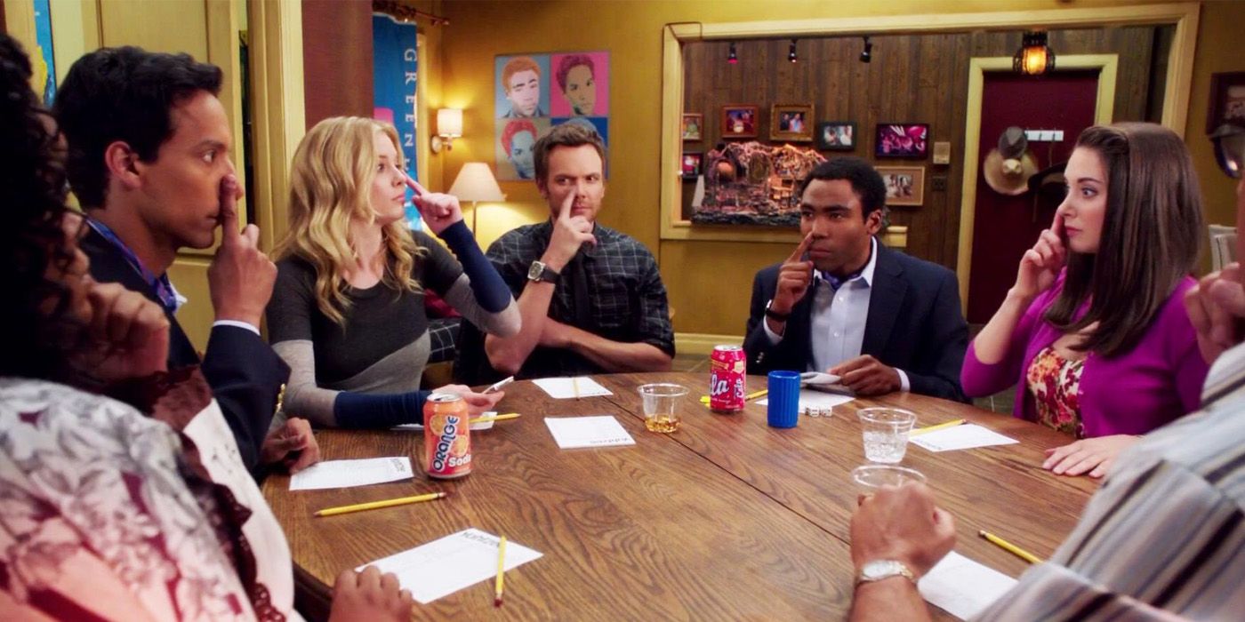 The characters of Community sit around a table and play Nose Goes
