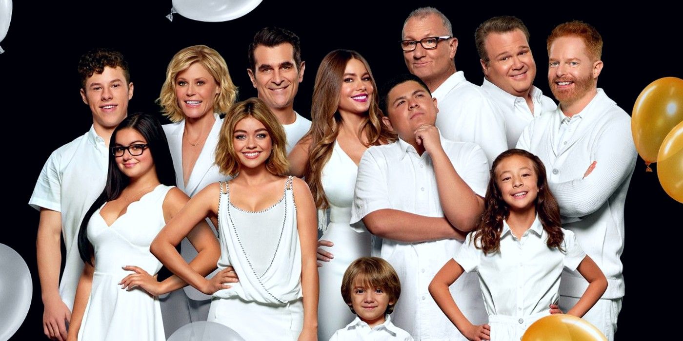 The cast of Modern Family stand in a group shot in white, in front of a black background, with balloons.