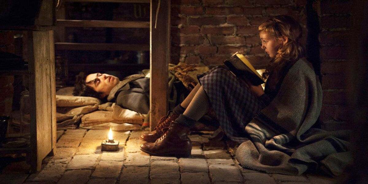 A girl reads a book by a sleeping man in The Book Thief