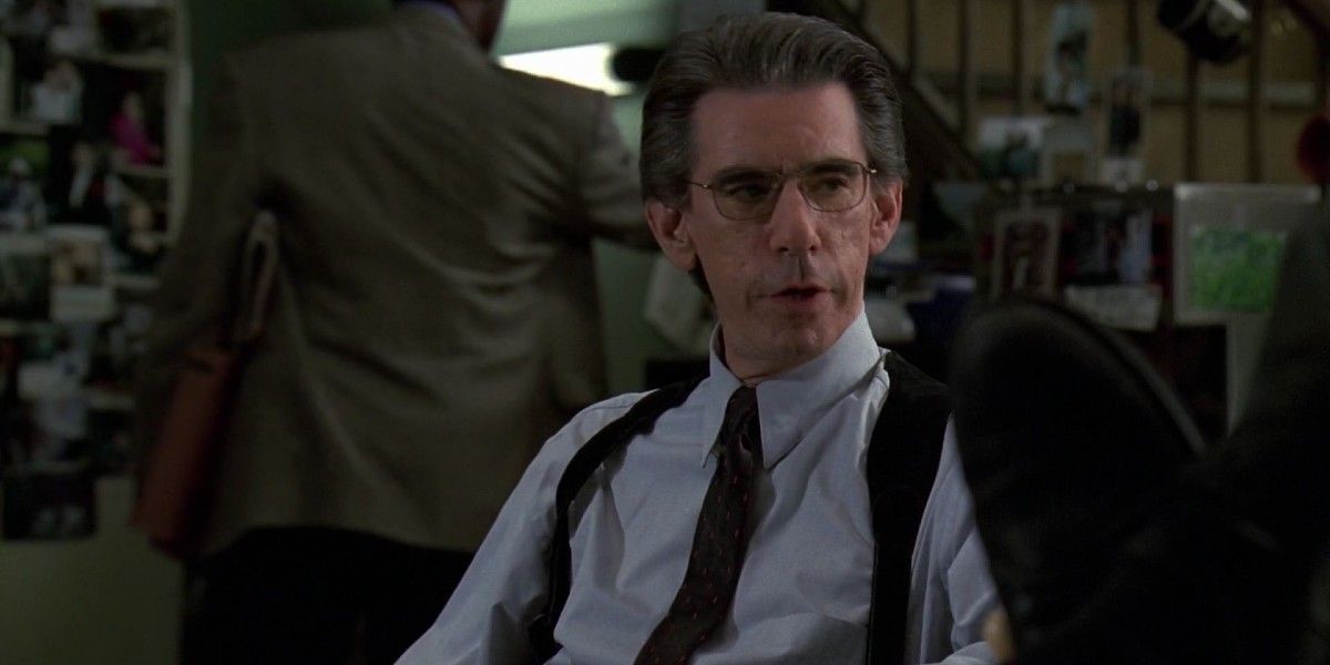 Detective Munch sits on his desk without his coat while talking to another detective in SVU