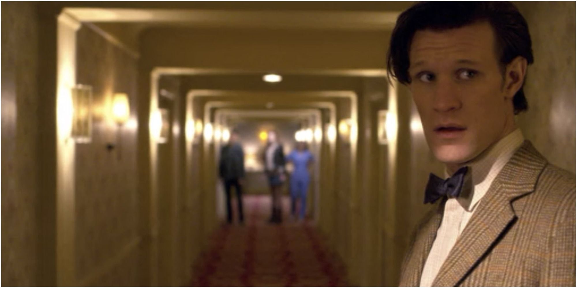 The Eleventh Doctor stands in a hotel hallway with others in the background behind him in Doctor Who.