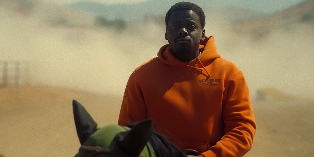Daniel Kaluuya riding horseback in an orange hoodie, with a dessert in the background as seen in the film Nope