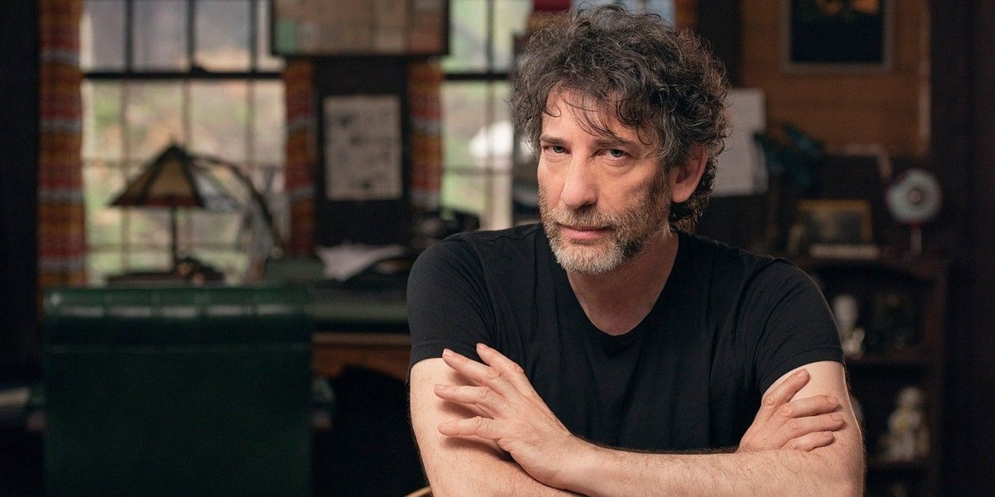 Neil Gaiman with his arms crossed, looking directly at the camera with his head tilted slightly, sitting in what looks like a home office