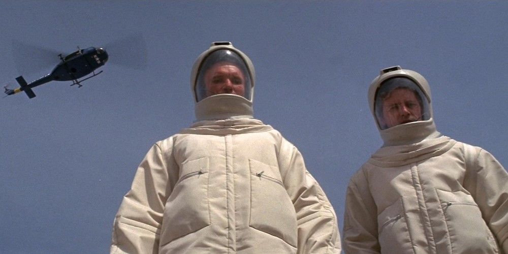 Two scientists in white hazmat suits look down the camera as a helicopter flies above.