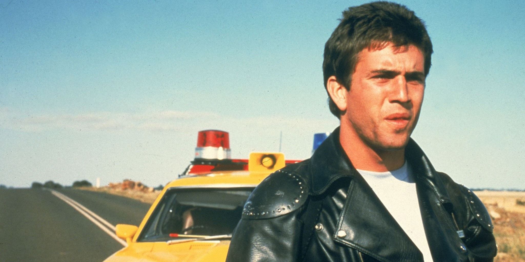 Mel Gibson in a black leather jacket and white tee standing behind his yellow police car.
