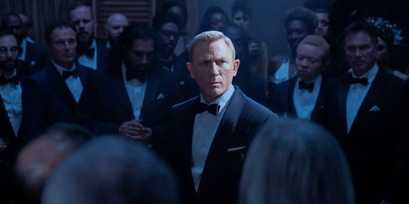 James Bond in a suit, surrounded by others, a light shining on him in a scene from No Time to Die.