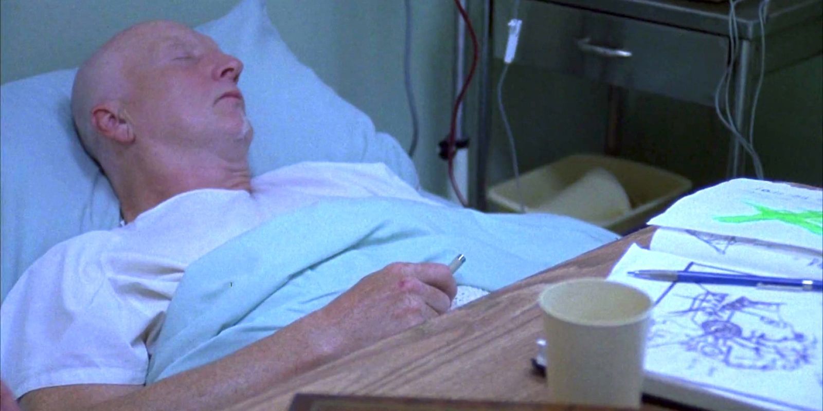 John Kramer, interpretado por Tobin Bell, em Saw sentado na cama do hospital segurando uma caneta.