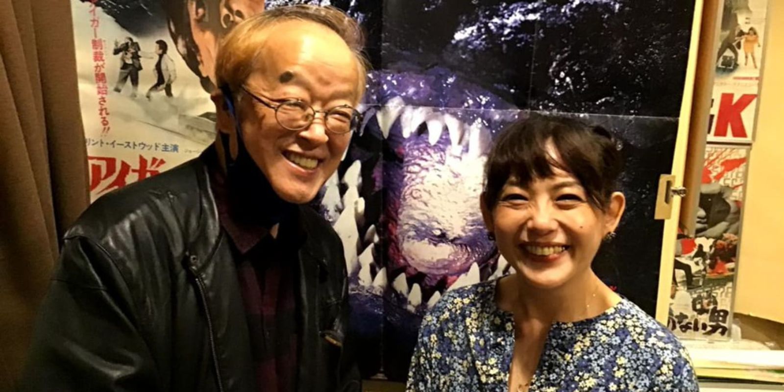 Kazuki Omori standing and smiling in front of a Godzilla poster
