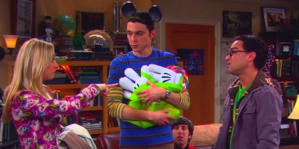 Sheldon wears Mickey Mouse ears and holds Disneyland paraphernalia as Leonard and Penny stand to either side of him - The Big Bang Theory