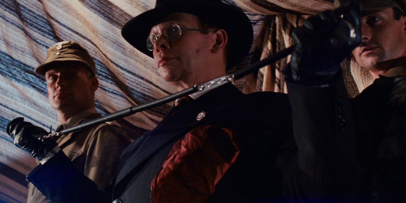 Ronald Lacey as Major Toht in Raiders of the Lost Ark. He is flanked by two soldiers in a tent as he assembles a coat hanger.