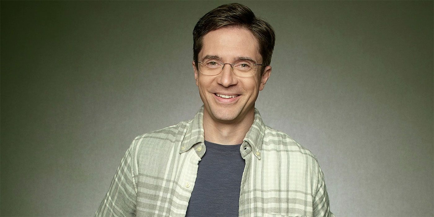 Topher Grace smiles in a plaid shirt against a grey background