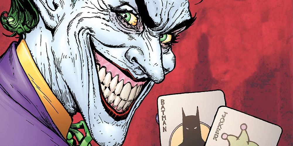 The Joker smiles and holds two playing cards - one depicting Batman and the other a Joker card.