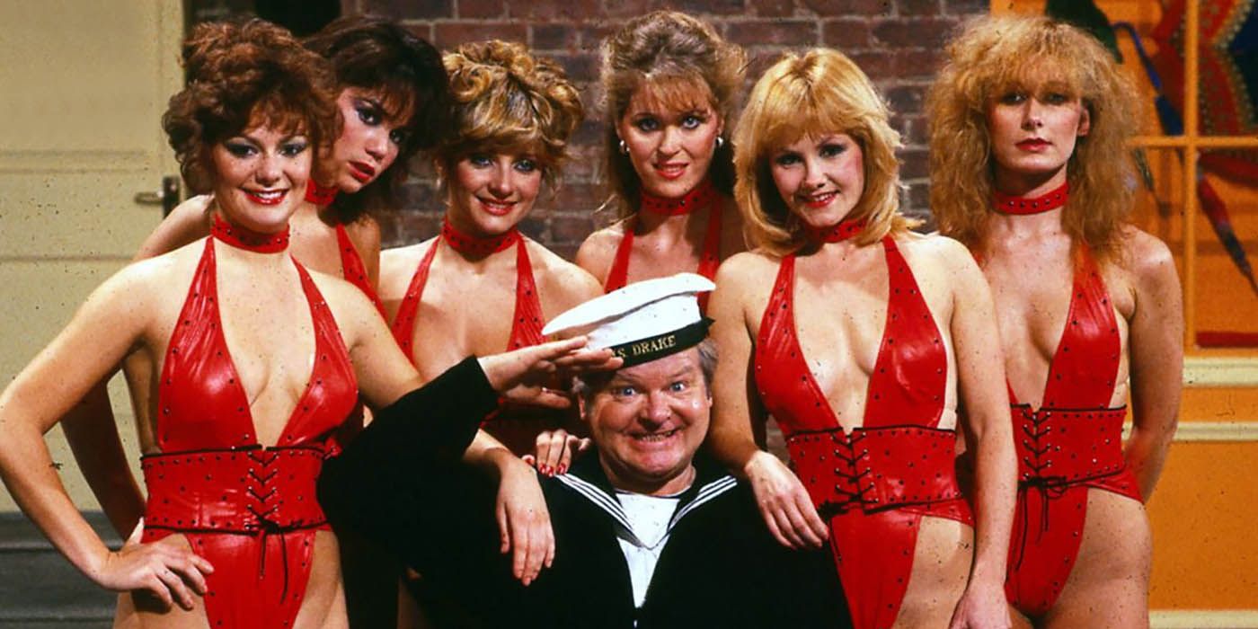 Benny Hill smiling in a sailor's uniform, saluting and surrounded by women in red bathing suits on The Benny Hill Show.