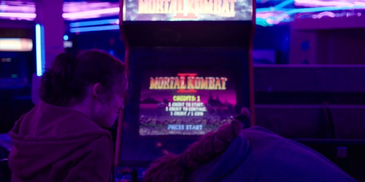Riley inserts coins into a Mortal Kombat II arcade cabinet as Ellie eaglerly awaits to play in episode seven in HBO's The Last of Us