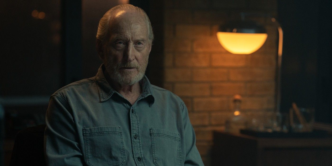 Charles Dance wearing a denim button up shirt sits in a dimly lit room.