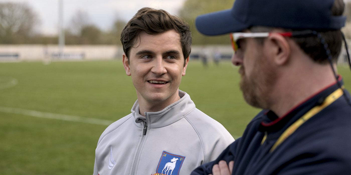 Colin de Ted Lasso sorrindo e conversando com o treinador Beard no campo.