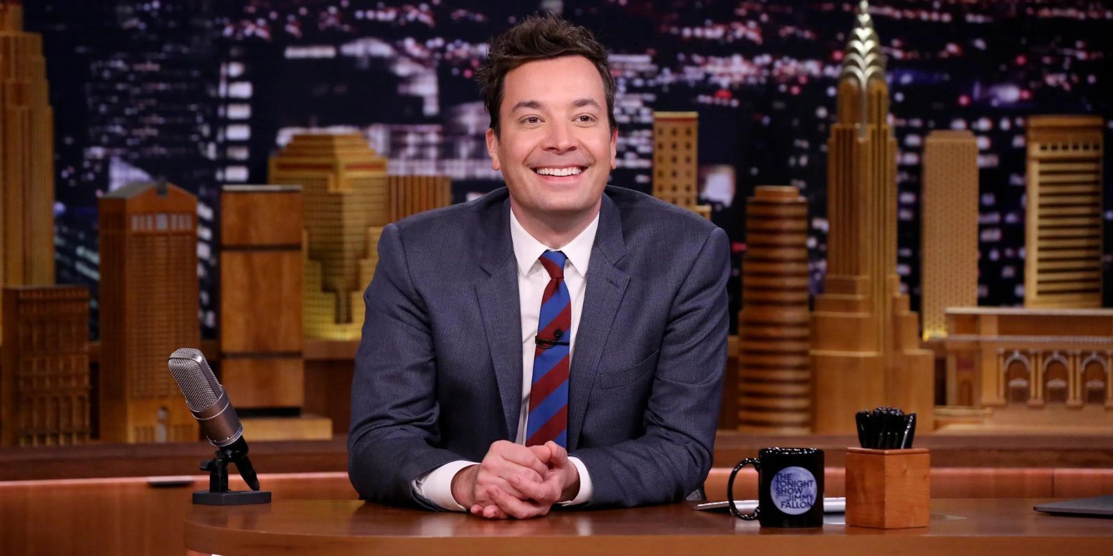 Jimmy Fallon sitting at his desk, smiling with his hands folded
