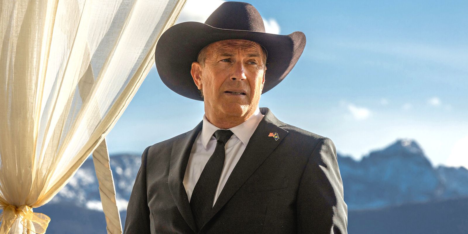 Kevin Costner in a black suit an ten-gallon hat, standing in front of a mountain range