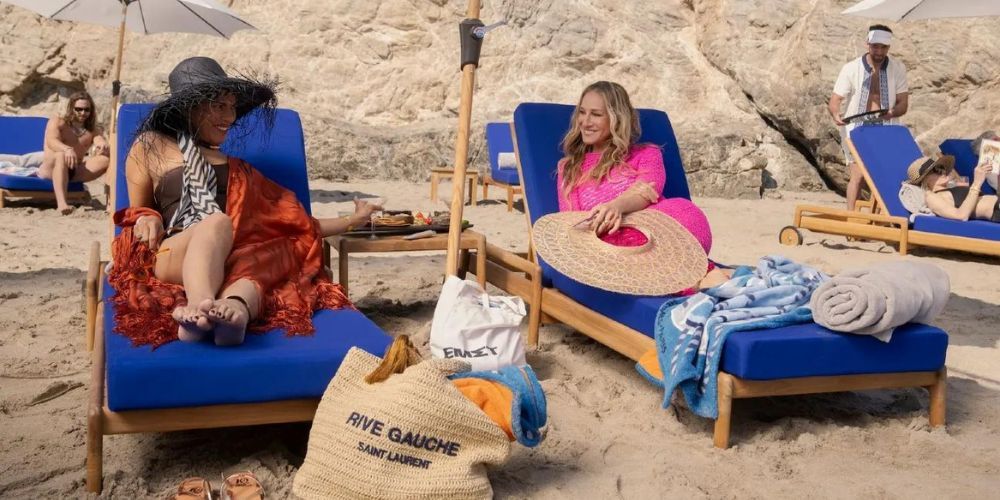 Sarita Choudhury como Seema Patel em um maiô preto com colcha vermelha, sentada em uma espreguiçadeira azul em uma praia ao lado de Sarah Jessica Parker como Carrie Bradshaw em rosa, que está sentada em uma espreguiçadeira azul em And Just Like That. ...