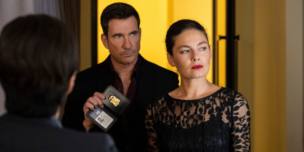 Alexa Davalos as Kristin Gaines and Dylan McDermott as Remy Scott holding his FBI badge stand in front of a yellow wall on FBI: Most Wanted