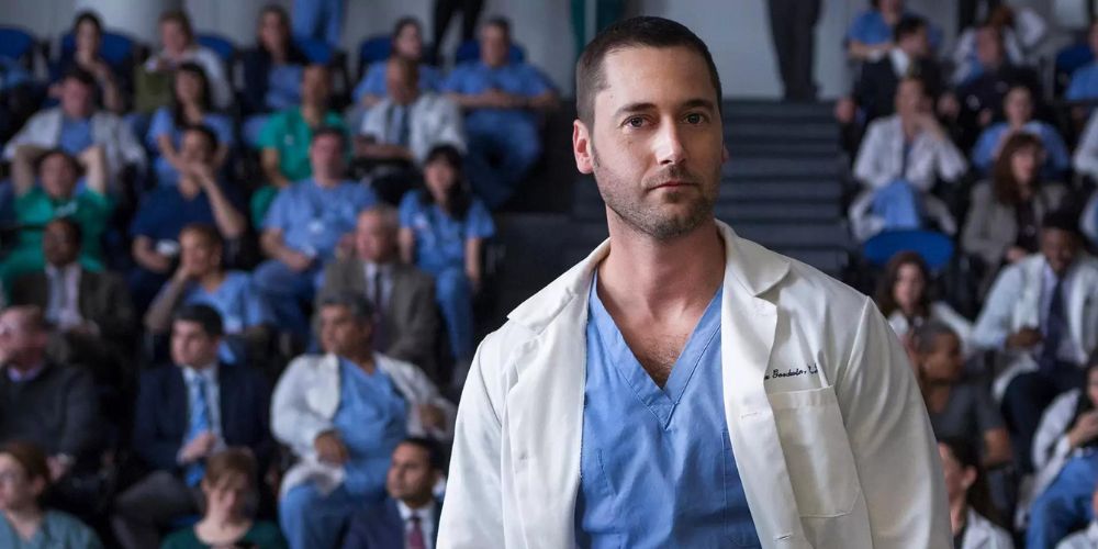 Ryan Eggold como Max Goodwin de uniforme médico e jaleco branco fica em frente a uma sala de funcionários de um hospital em New Amsterdam.
