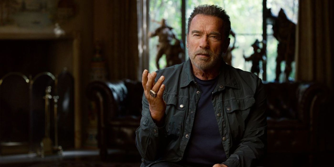 Arnold Schwarzenegger sitting in a chair holding his hand out while talking in a scene from Arnold.