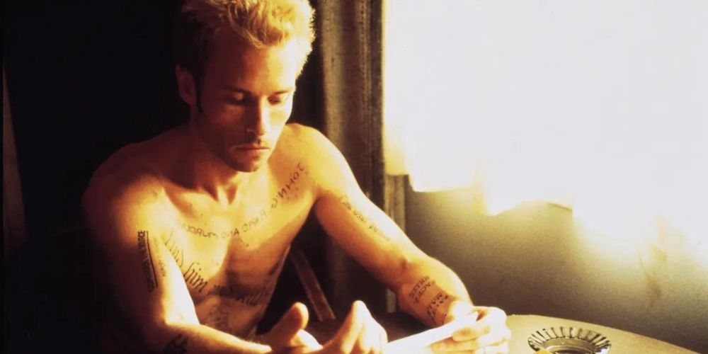 Guy Pearce as Leonard sits at a table with no shirt on, covered in tattoos, in Memento