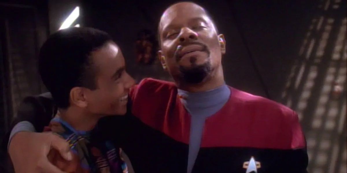 Avery Brooks, a bald Black man with a goatee, as Captain Sisko, embracing a smiling Cirroc Lofton, a young Black man with close-cropped black hair, who played Sisko's son Jake. 
