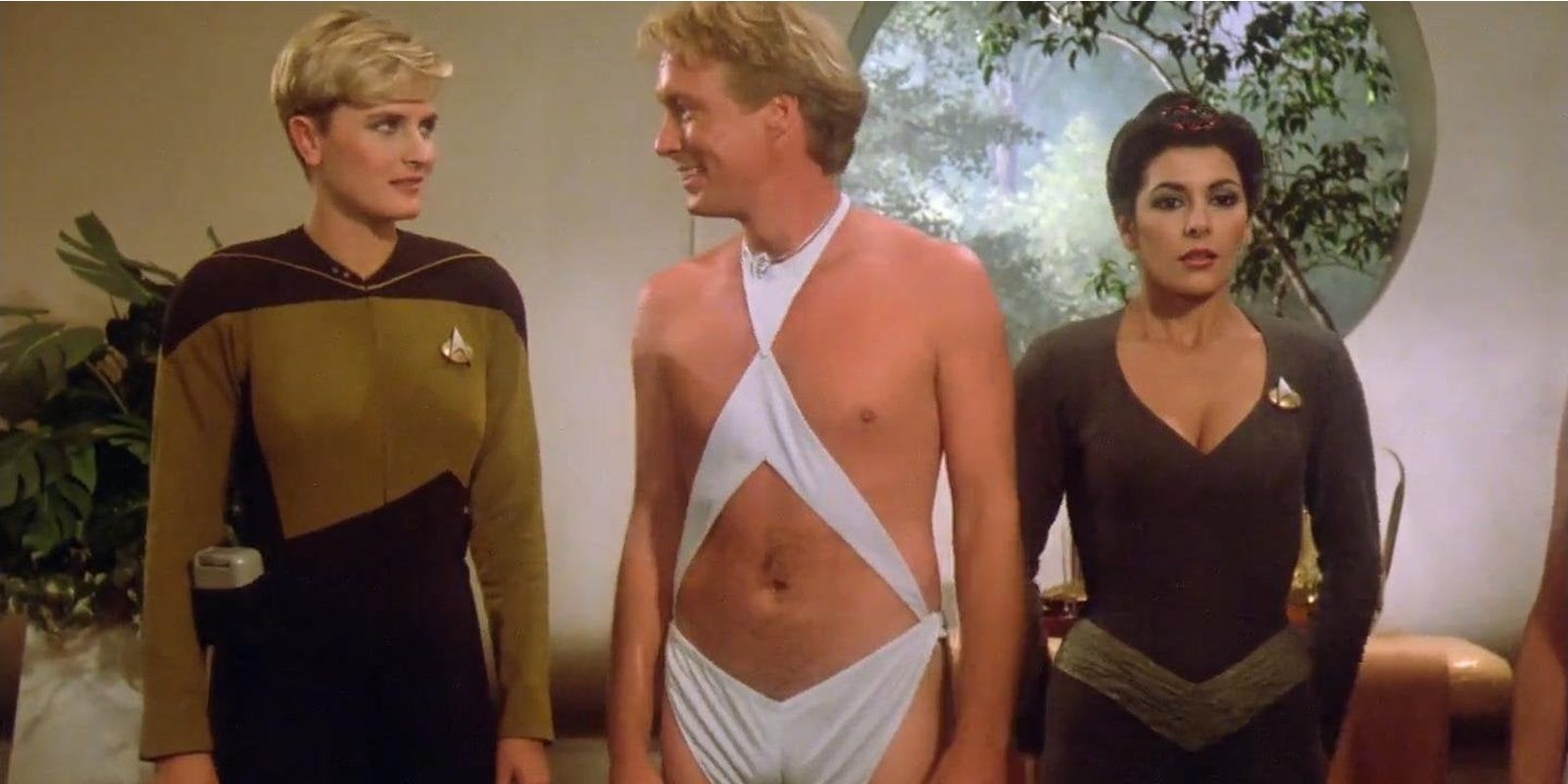 Tasha Yar e Deanna Troi do outro lado de um homem com uma roupa branca reveladora de Star Trek TNG