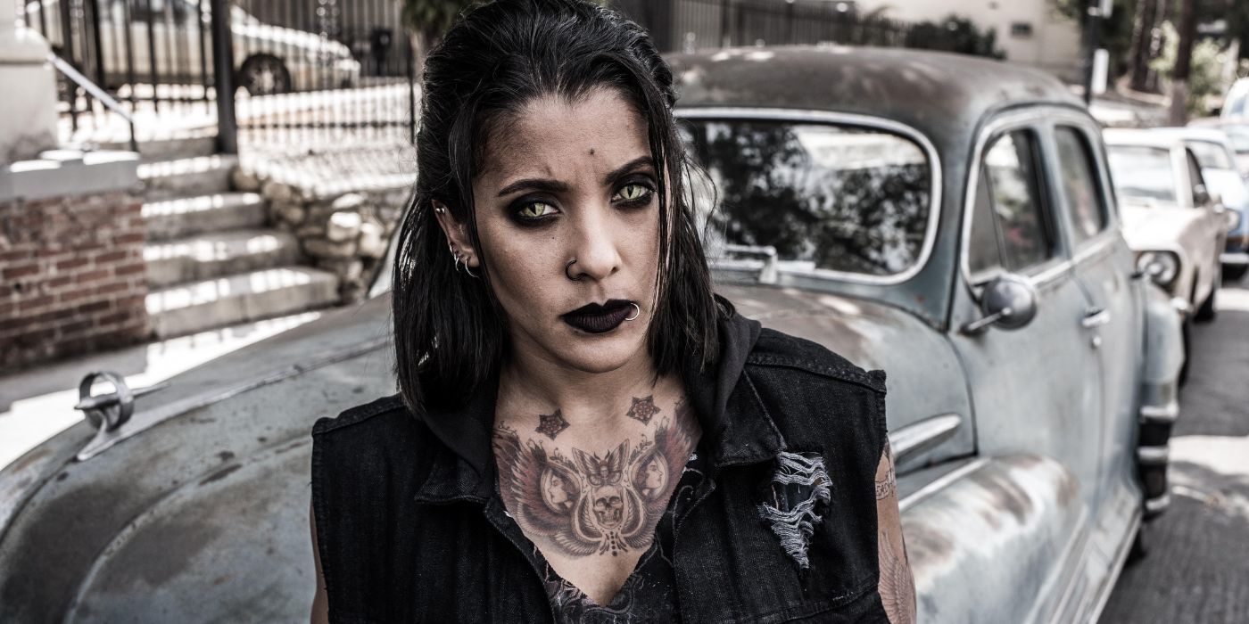 Desi Gutierrez looking at the camera with snake eyes, standing in front of a rusty car. 
