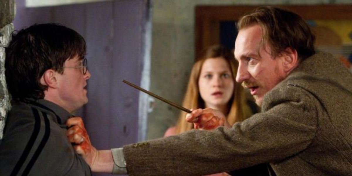 Remus Lupin holds Harry Potter against a wall and points his wand at him. Ginny in the background.