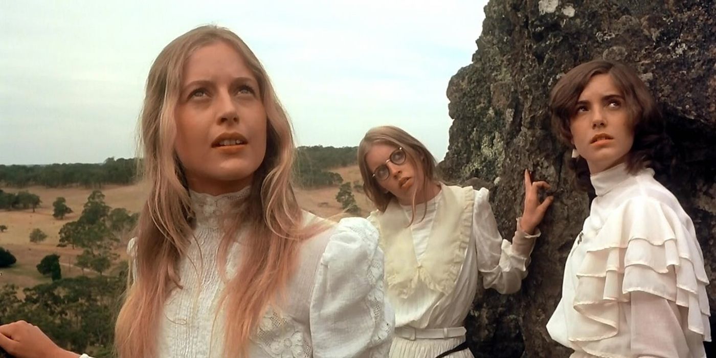 Three girls standing by a rock wearing white dresses and looking up at something in Picnic at Hanging Rock.