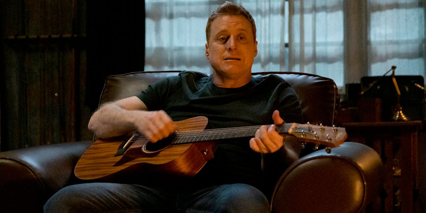 Resident Alien's Harry (actor Alan Tudyk) plays a guitar seated in black T-shirt