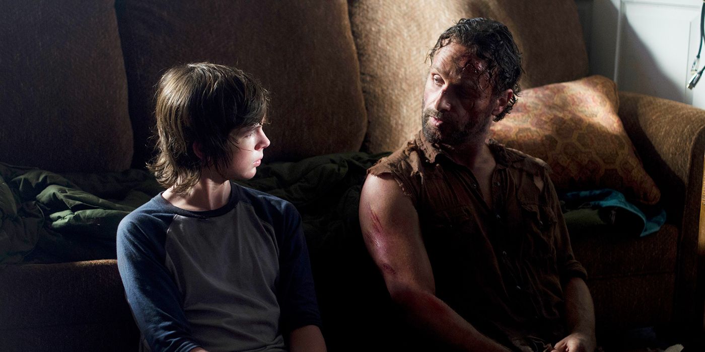 Carl and a battered Rick sitting on the floor in a house talking in The Walking Dead.