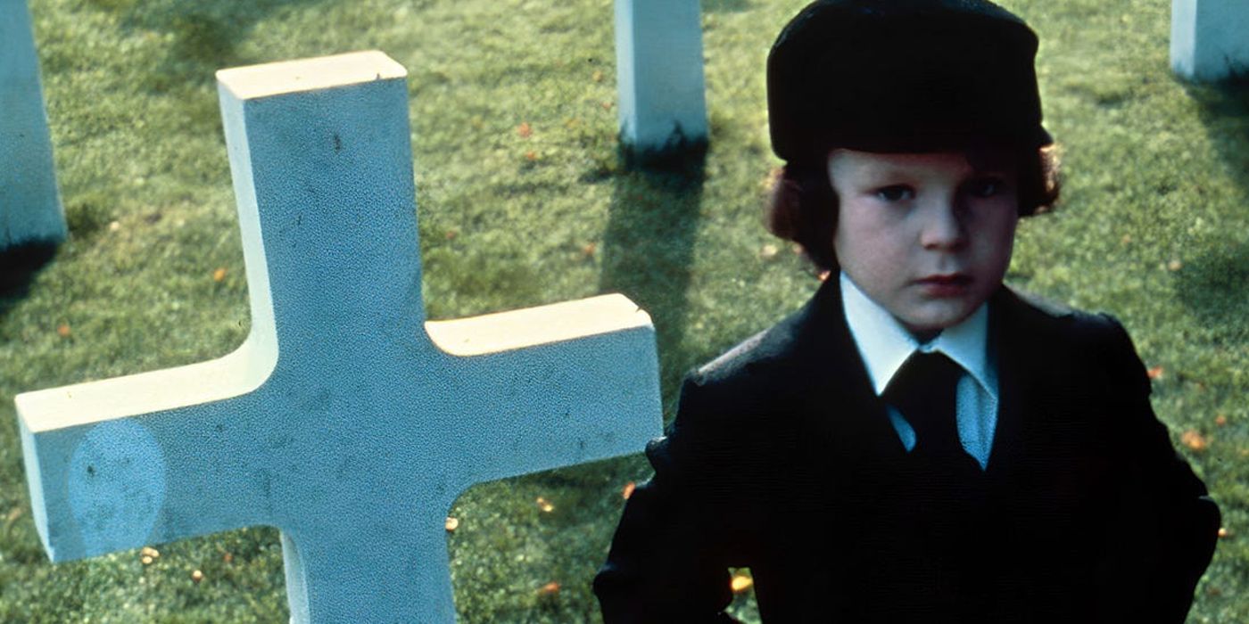 Damien stands emotionless in front of a cemetery cross in The Omen