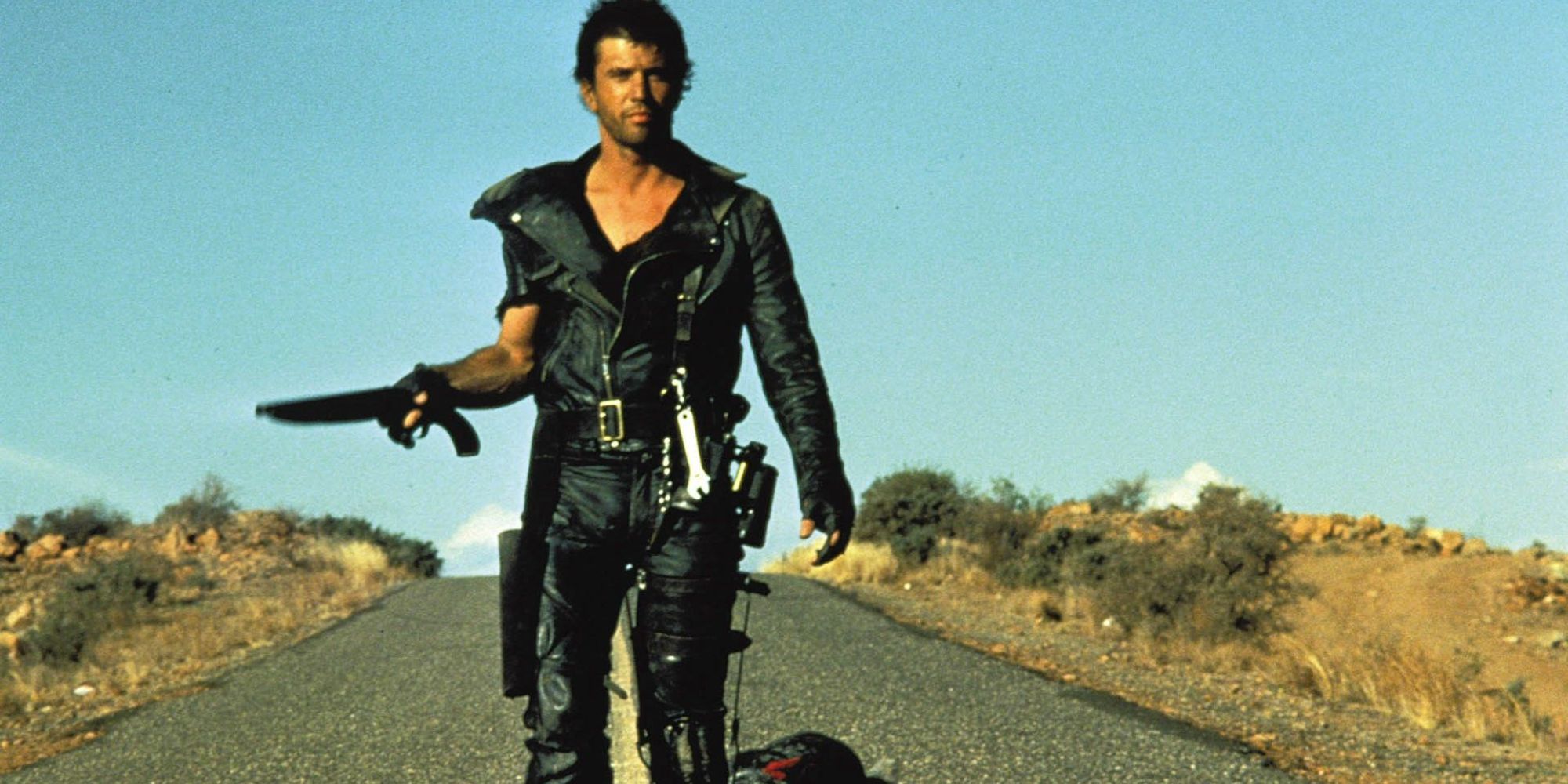 Mel Gibson walks down the road as Mad Max with a shotgun in The Road Warrior