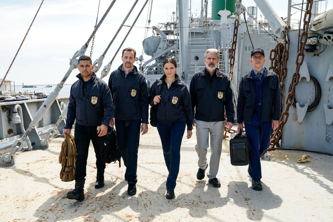 The entire NCIS team walks toward camera in NCIS gear on a ship's deck