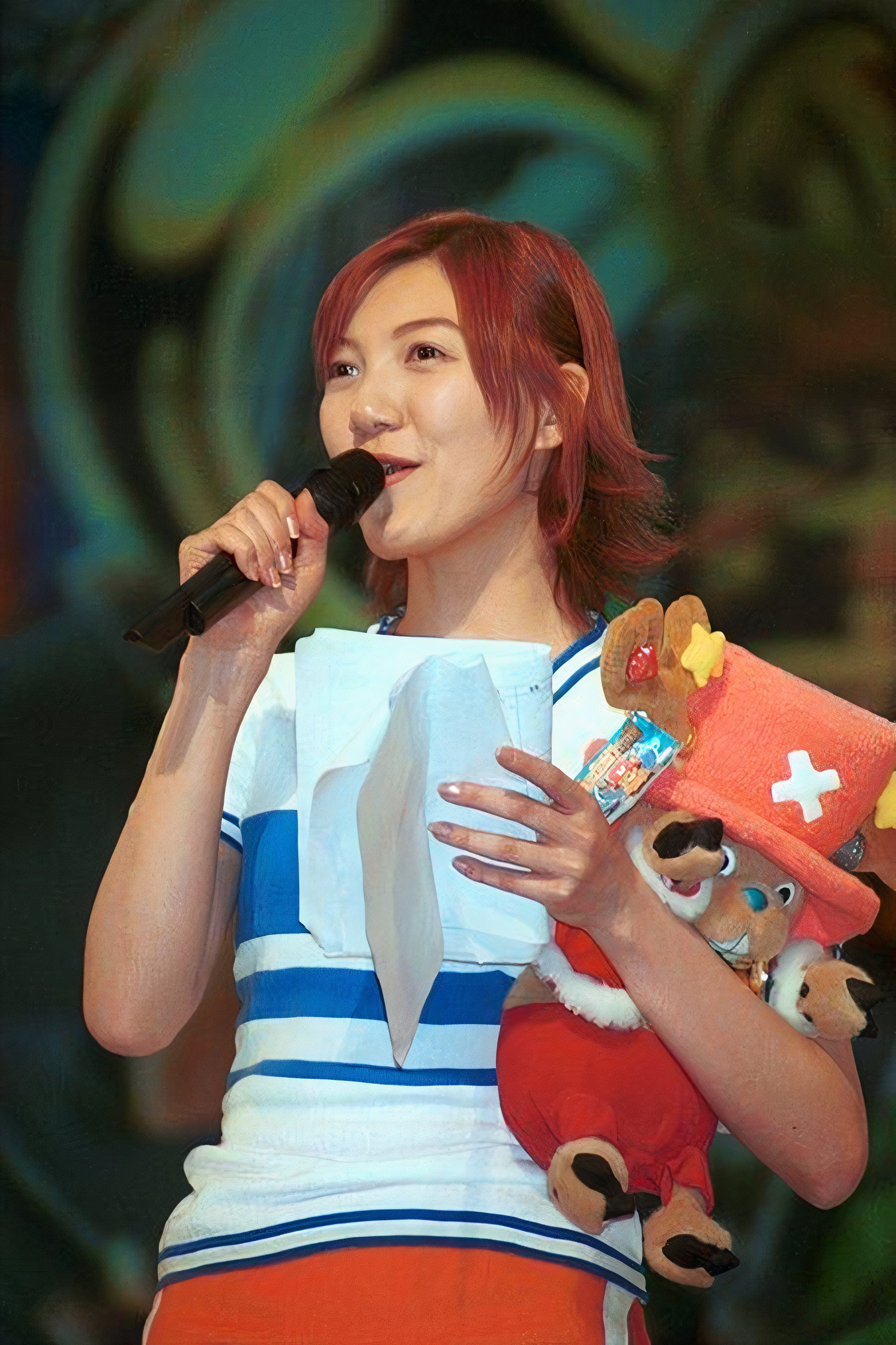 Chiaki Inaba is dressed as Nami and holding a Chopper stuffed animal while speaking into a microphone.