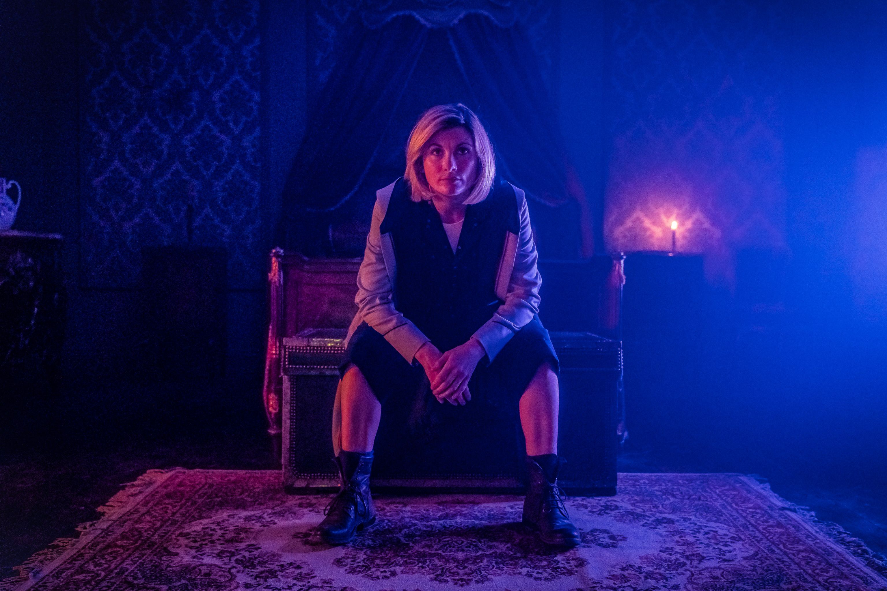 The Thirteenth Doctor sitting in a chair in a blue and pink lit room from Doctor Who