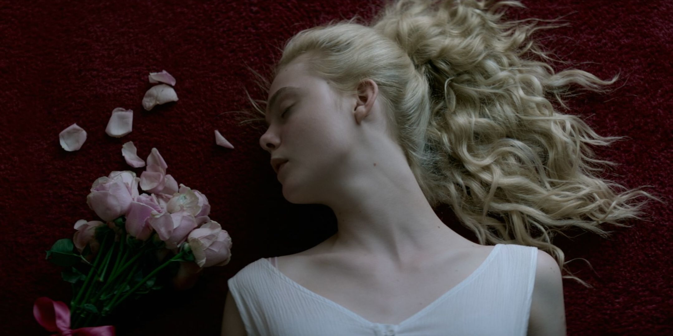 Elle Fanning as Jesse in The Neon Demon lays with her blonde hair in a pony tail on red carpet with flowers scattered next to her