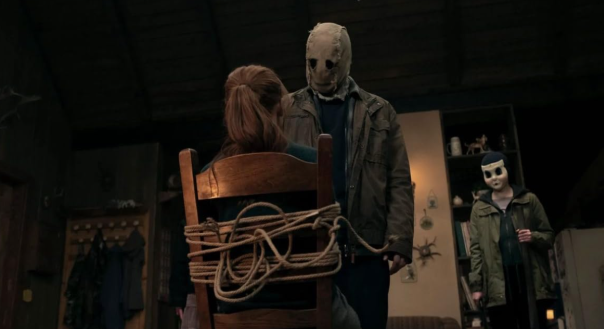 Maya está amarrada a uma cadeira com o Espantalho em pé sobre ela em The Strangers: Chapter 1.