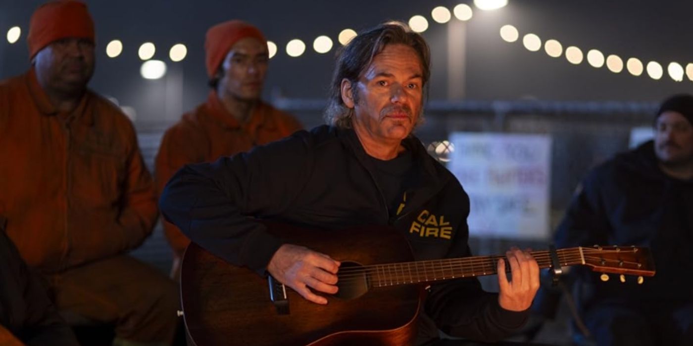 Billy Burke as Vince Leone plays the guitar in front of a fire on Fire Country