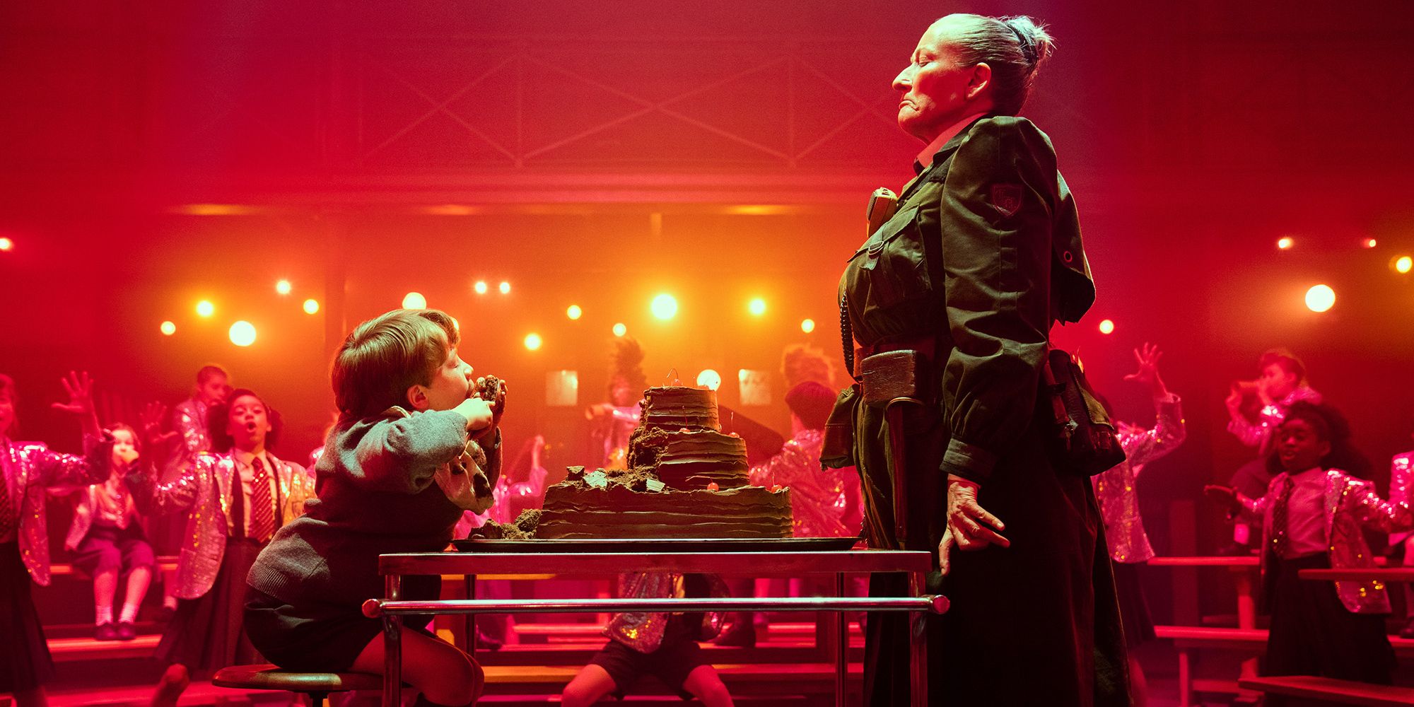 Sra. Trunchbull força Bruce a comer um bolo inteiro enquanto os alunos cantam e dançam ao fundo para animá-lo em Matilda the Musical