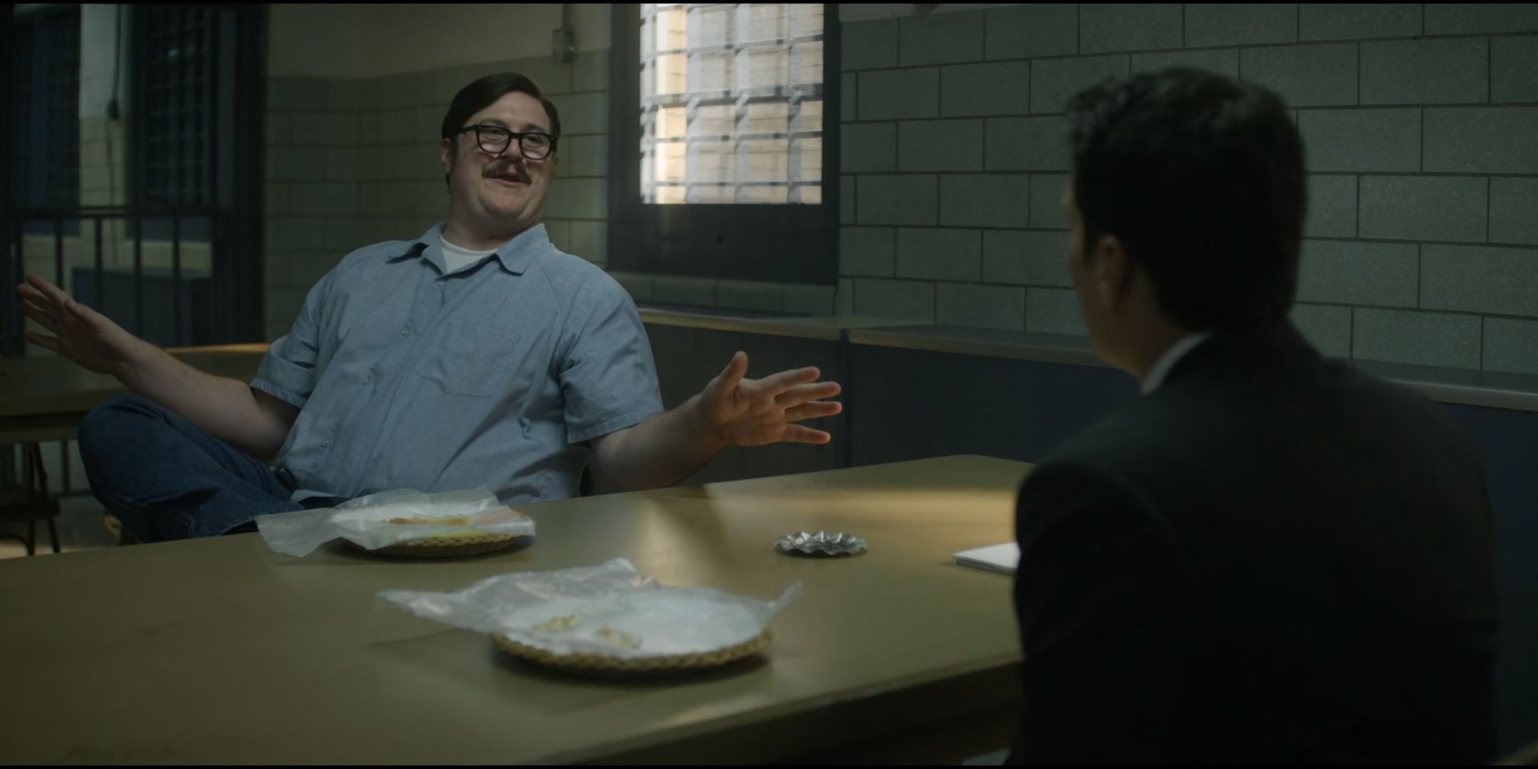 Cameron Britton as Ed Kemper spreads his arms and smiles at a table as he talks to Jonathan Groff as Holden Ford in Mindhunter